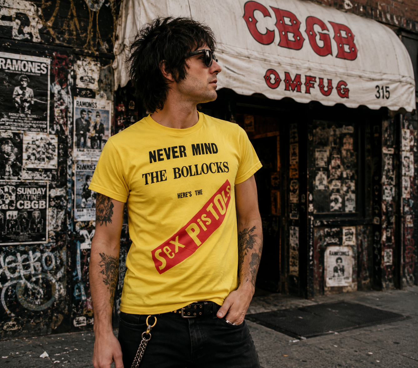 Male model wearing a yellow Sex Pistols Never Mind The Bollocks T-shirt outside a graffiti-covered punk rock club, showcasing iconic British punk style and classic album artwork from a side-angle view.