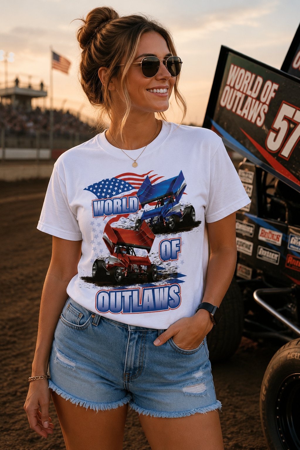 Female model wearing the World of Outlaws American Flag White T-Shirt at a dirt track racing venue during sunset, featuring red and blue sprint car graphics, patriotic American flag artwork, and a sprint car in the background.