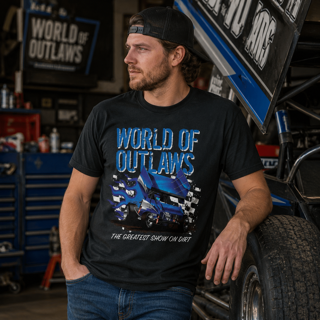 Male model wearing a World of Outlaws Blue Fire Black T-Shirt while standing beside a sprint car inside a racing garage workshop