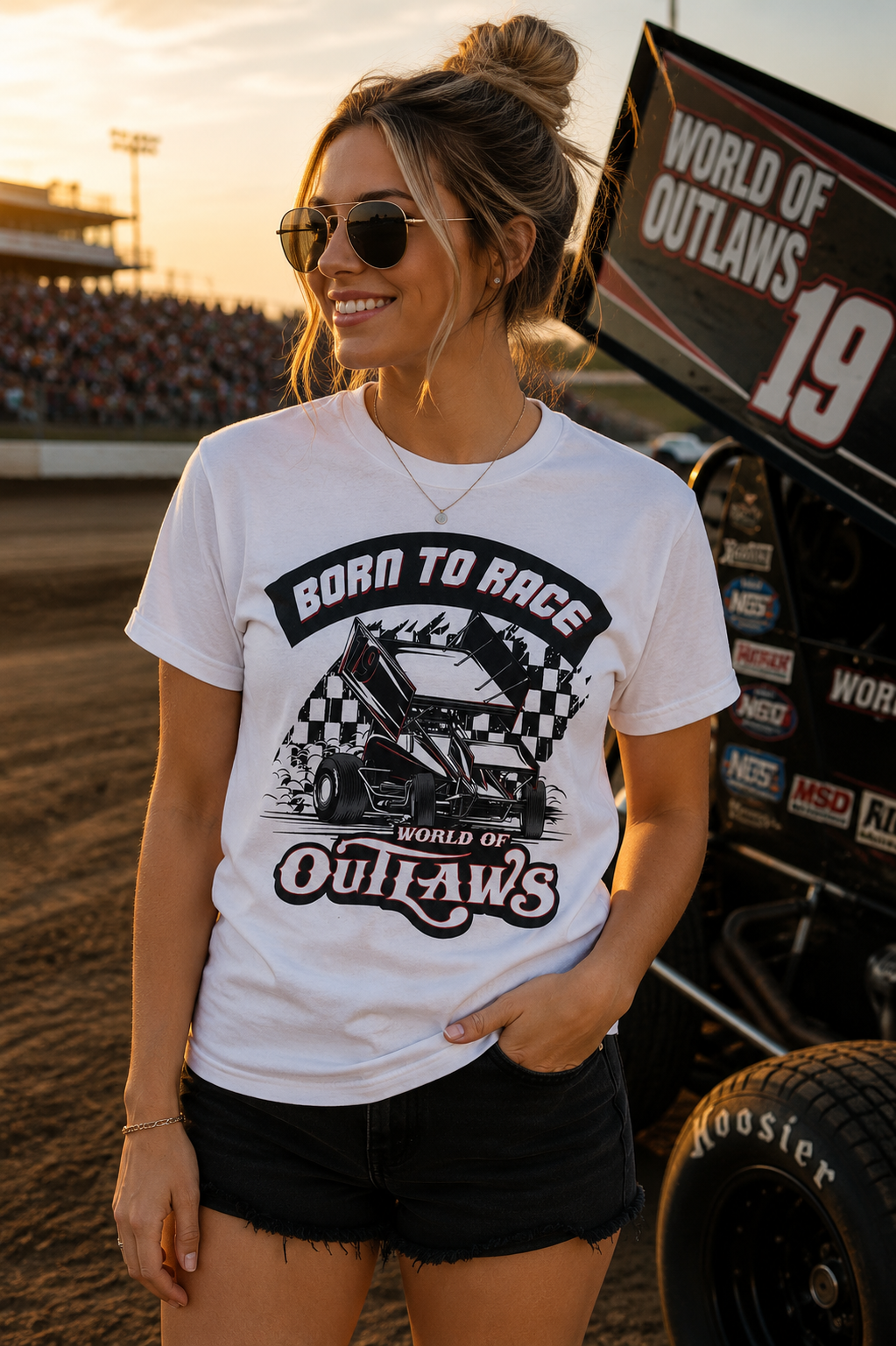 Female model wearing the World of Outlaws Born To Race Black Flag White T-Shirt at a dirt track racing venue during sunset, featuring a vintage sprint car graphic, checkered flag artwork, and World of Outlaws racing design with a sprint car and grandstands in the background.