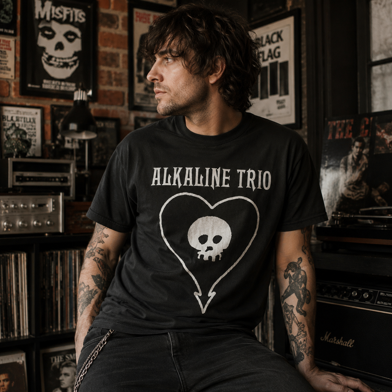 Male model wearing an Alkaline Trio Classic Heartskull black t-shirt in a vintage rock music room with records, posters, and guitar amps.