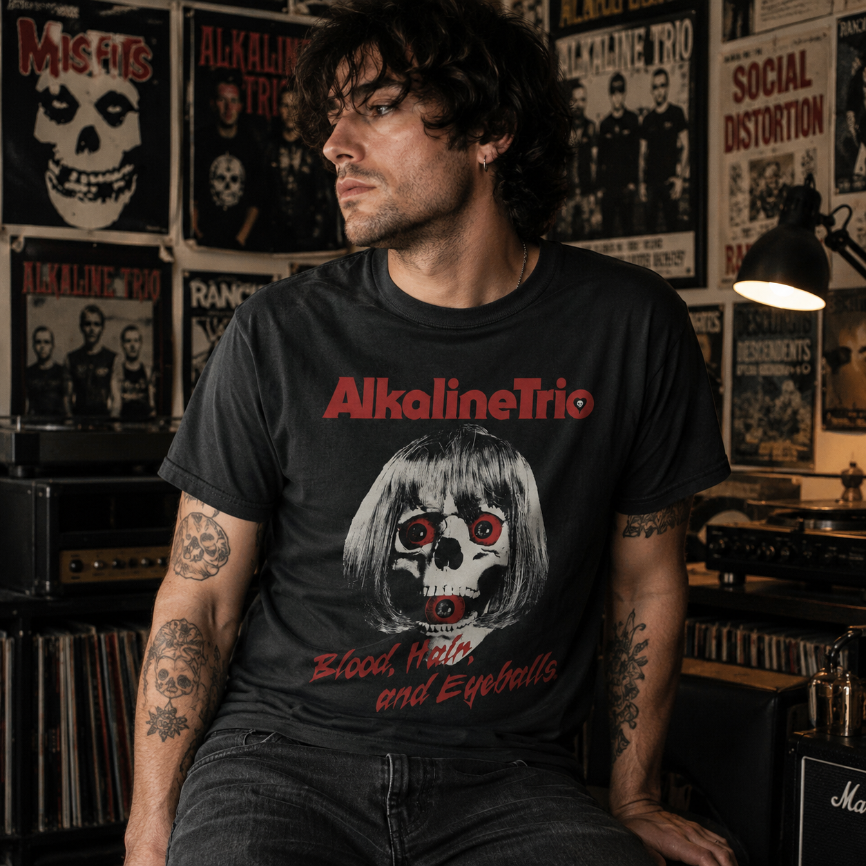 Male model wearing an Alkaline Trio Skelly Eyes black t-shirt in a moody punk rock music room with posters, records, and guitar amps.