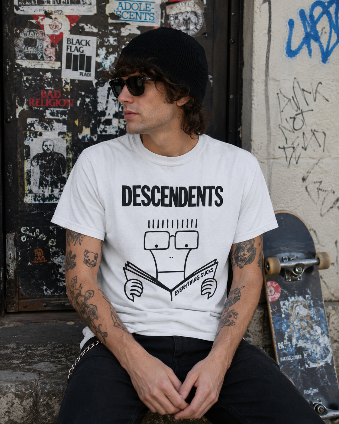 Male model wearing white Descendents Everything Sucks punk band t-shirt in urban skateboarding street setting with graffiti background and skateboard.