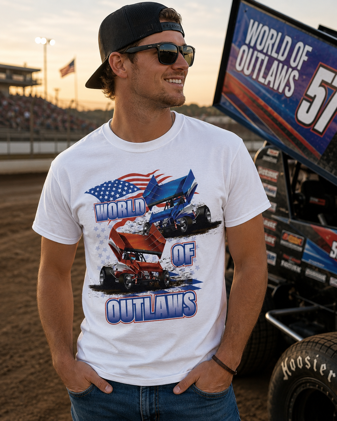 Male model wearing the World of Outlaws American Flag White T-Shirt at a dirt track racing venue during sunset, featuring red and blue sprint car graphics with patriotic American flag artwork and a sprint car in the background.