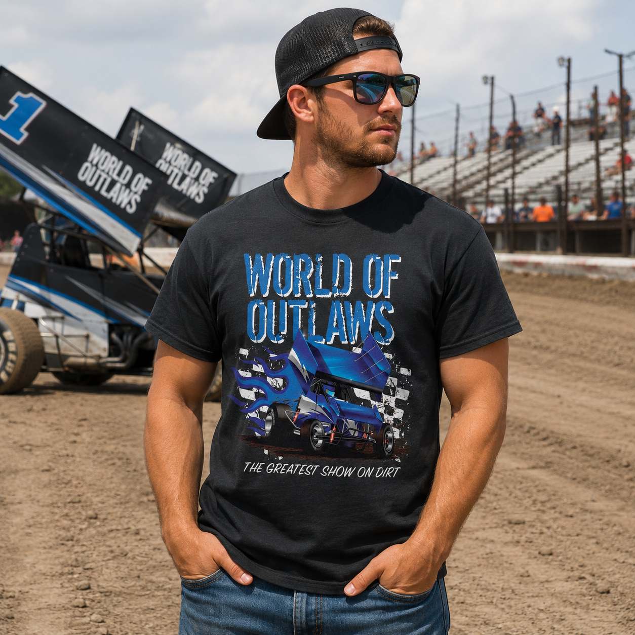 Male model wearing a World of Outlaws Blue Fire Black T-Shirt at a dirt track racetrack with sprint car and grandstands in the background