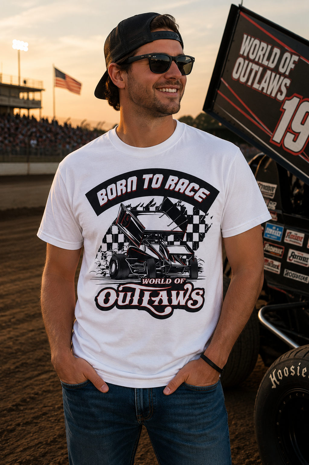 Male model wearing the World of Outlaws Born To Race Black Flag White T-Shirt at a dirt track racing venue during sunset, featuring a vintage sprint car graphic, checkered flag artwork, and World of Outlaws racing design with a sprint car in the background.