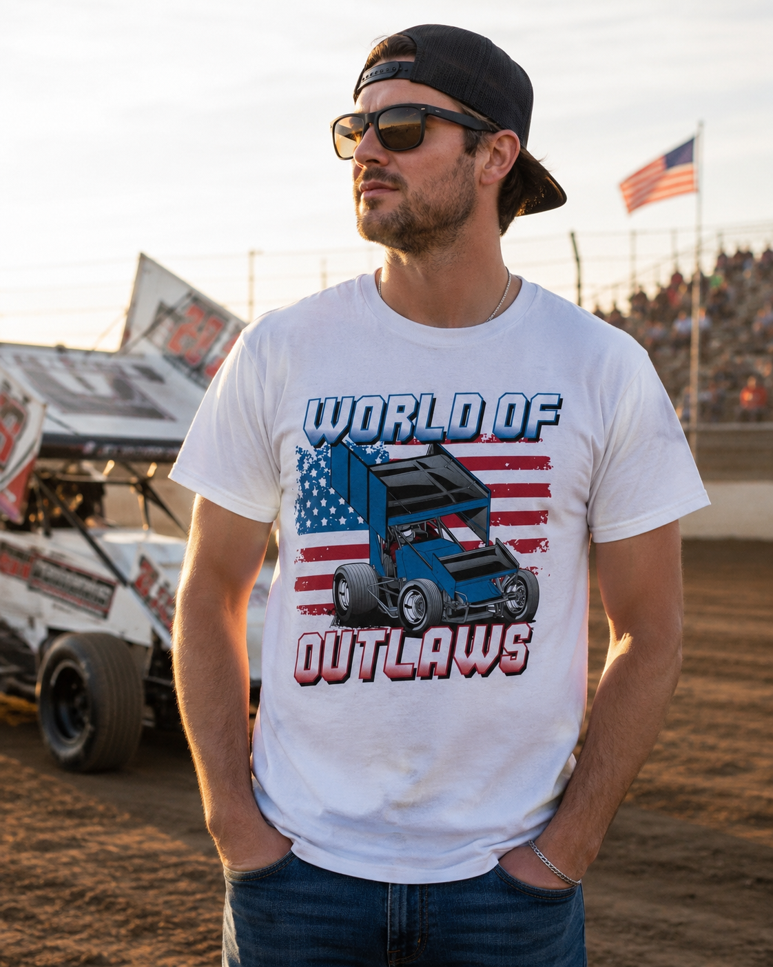 Male model wearing a World of Outlaws Gradient American Flag White T-Shirt at a dirt track raceway with sprint cars and grandstands in the background during sunset.