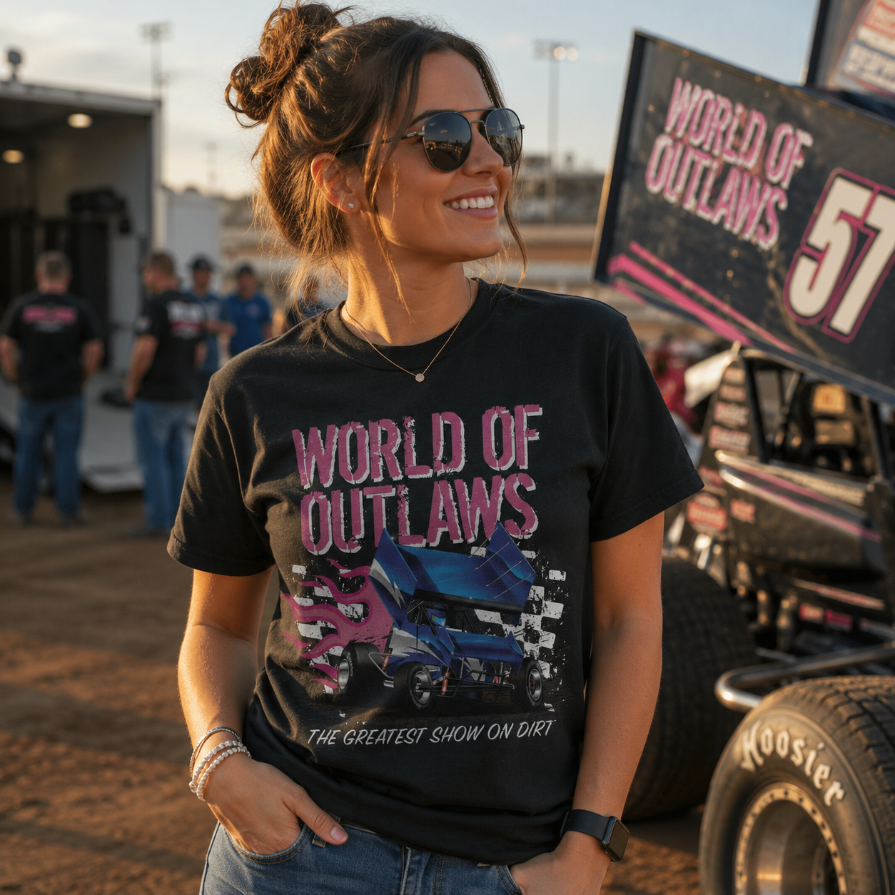 Female model wearing the World of Outlaws Grunge Logo Black T-Shirt at a dirt track racing venue with a sprint car in the background during golden hour, showcasing the distressed sprint car graphic and Greatest Show on Dirt design.