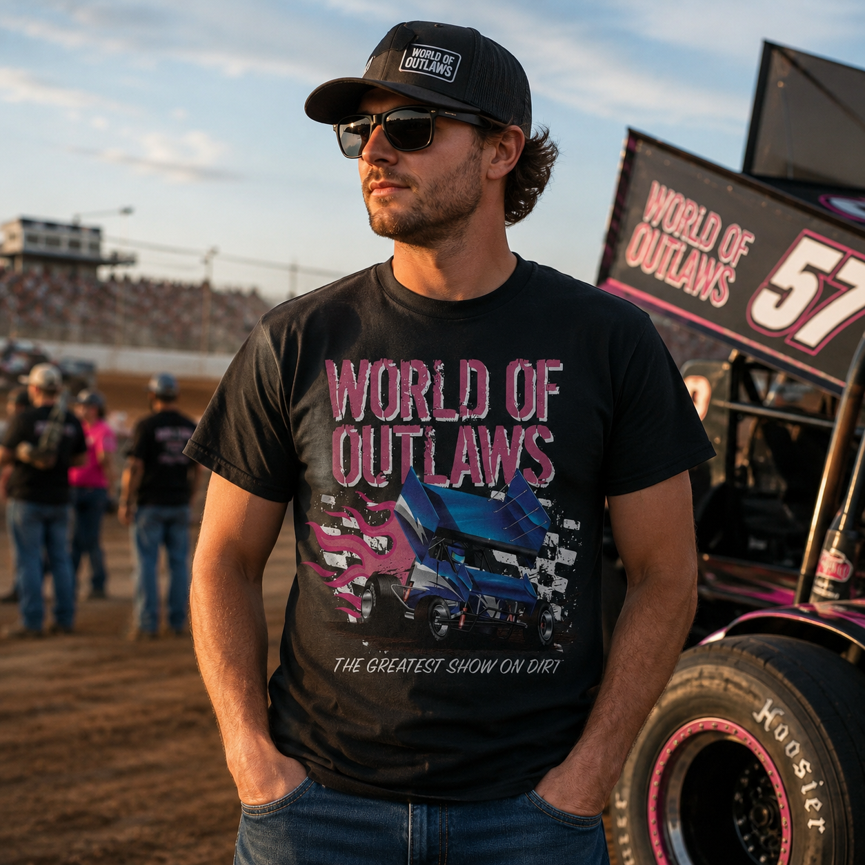 Male model wearing the World of Outlaws Grunge Logo Black T-Shirt at a dirt track racetrack with sprint car and grandstands in the background during golden hour.