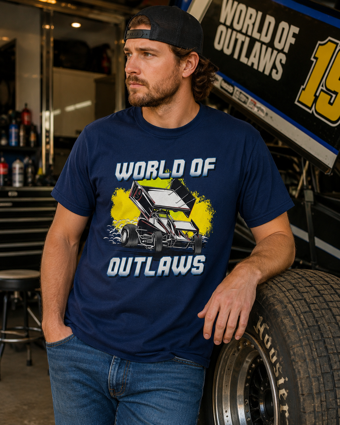 Male model wearing a World of Outlaws Yellow Splash Navy T-Shirt inside a sprint car racing garage with race car, tires, and workshop equipment in the background