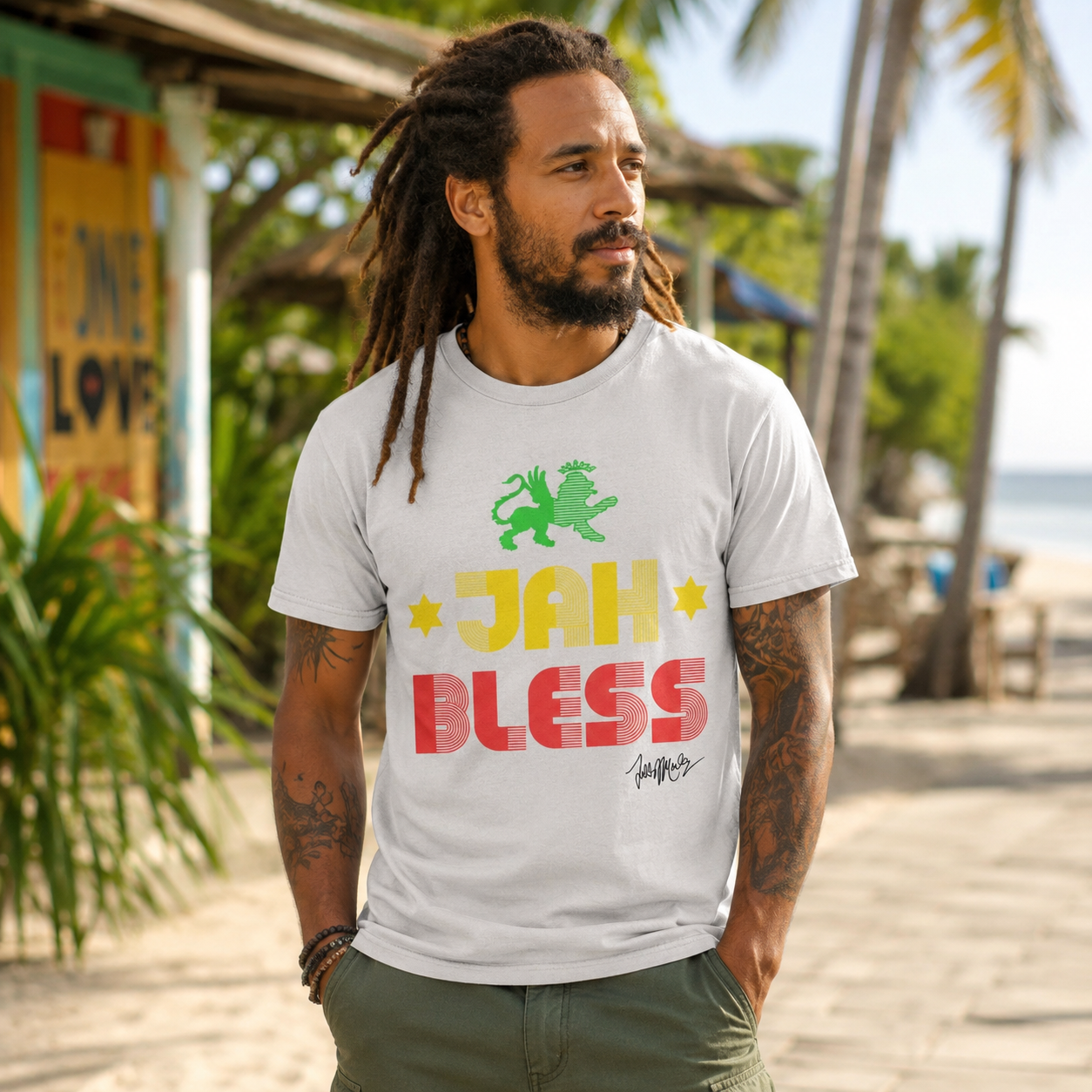 Male model wearing a Ziggy Marley Jah Bless Ice Grey T-Shirt on a tropical beach walkway with palm trees, reggae-inspired surroundings, and ocean views in the background.