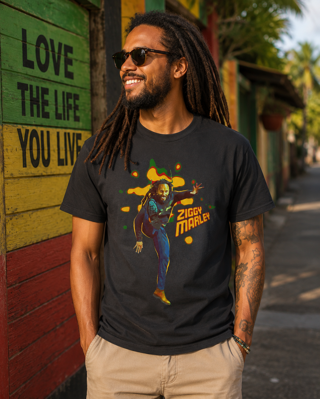Male model wearing the Ziggy Marley Kick Ziggy black t-shirt in a colorful reggae-inspired street setting with tropical surroundings, sunglasses, and vibrant mural artwork in the background.